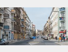Straßenszene in einer urbanen Wohngegend mit mehrstöckigen Altbauhäusern auf beiden Seiten der Straße, die über Balkone und teils verzierte Fassaden verfügen. Am Straßenrand parken Autos, während einige Menschen auf den Gehwegen unterwegs sind; ein Verkehrsschild zur Richtungsführung steht in der Mitte der Fahrbahn.