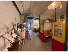 Ein industriell gestalteter Innenraum mit weiß gestrichenen Backsteinwänden, gewölbter Decke und sichtbaren Lampen, die an einer Holzleiste montiert sind. Rechts steht eine gelb-rot gestrichene alte Schienenfahrzeugkabine mit Holzbänken, während der Raum im Hintergrund einen Arbeitsplatz und weitere Gegenstände zeigt.