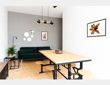 Heller, moderner Wohnraum mit einem großen Holztisch und integriertem Metallgestell samt runden Sitzen, einer grünen Couch vor einer grauen Wand und minimalistischer Wanddekoration, darunter Spiegel und Kunstwerke. Holzboden und eine Zimmerpflanze auf einem Beistelltisch runden das Design ab.