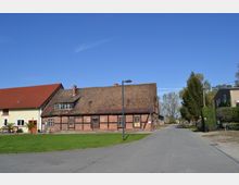 Ein Dorfplatz mit einem traditionellen Fachwerkgebäude aus rotem Backstein und einem angrenzenden gelb verputzten Haus mit roten Dächern. Im Vordergrund ist eine asphaltierte Straße, im Hintergrund sind Bäume und einzelne Häuser sichtbar.