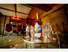 Eine Nahaufnahme einer Whiskyflasche der Marke "Stork Club" auf einem silbernen Tablett, umgeben von mehreren Gläsern. Im Hintergrund ist ein holzvertäfelter Raum mit Musikinstrumenten, einer Sitzgruppe und gedämpfter Beleuchtung erkennbar.