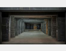 Industrieähnlicher Raum mit mehreren hintereinander angeordneten Betonrahmen, die einen langen, perspektivischen Tunnelblick formen. Die Wände und Böden wirken roh und unverkleidet, mit dunklem Beton und vereinzelten Holz- und Metallstrukturen im Hintergrund.