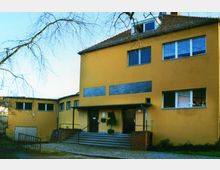 Gelbes Gebäude mit Flachdach- und Satteldach-Elementen, großer Treppenaufgang mit Metallgeländern, ziegelverkleideten Sockeln und mehreren Fenstern unterschiedlicher Größe. Im Vordergrund ist ein gepflasterter Hofbereich mit kahlen Bäumen sichtbar.