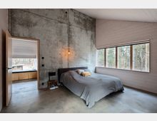 Minimalistisch eingerichtetes Schlafzimmer mit Betonwand, einem großen Bett mit grauer Tagesdecke und einer hängenden geometrischen Lampe. Auf der rechten Seite befindet sich ein großes Fenster mit Blick auf einen Wald, und links führt eine geöffnete Tür in einen angrenzenden Raum.
