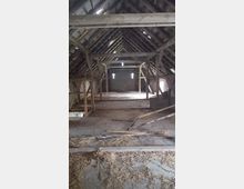 Alter Dachboden mit sichtbarer Holzkonstruktion, offenen Dachbalken und vereinzelten Lichteinfällen durch kleine Fenster in Kreuzform. Der Boden ist teilweise mit Stroh und Holzresten bedeckt.