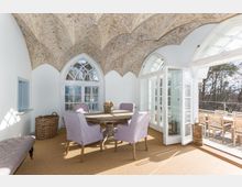 Esszimmer mit gewölbter, kunstvoll bemalter Decke, Rundbogenfenstern und einer Glastür, die zu einer Terrasse mit Holzgartenmöbeln führt. Im Raum stehen ein runder Holztisch und Stühle mit fliederfarbenem Stoffbezug.