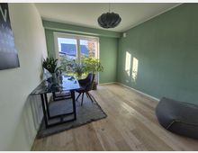 Minimalistisch eingerichtetes Arbeitszimmer mit grün gestrichenen Wänden, Holzfußboden und großem Fenster, durch das Tageslicht hereinfällt. Ein Schreibtisch aus Glas mit Pflanzen darauf und ein gepolsterter Stuhl stehen auf einem Teppich; rechts ein großer Sitzsack.