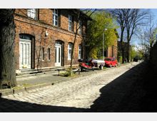 Gepflasterte Straße mit roten Backsteinhäusern auf der linken Seite, weißen Türen und Fenstern; ein geparkter Roller mit Anhänger und ein rotes Auto stehen am Straßenrand, umgeben von Bäumen und Laternen.