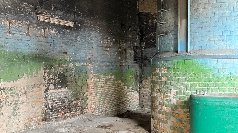 Innenraum eines alten Industriegebäudes mit abgenutzten, teils grün gestrichenen Ziegelwänden, Betonstufen und freiliegender Holzbalkendecke. Rechts steht ein großer zylindrischer gemauerter Tank mit Leiter sowie ein grüner Metallbehälter auf dem Betonboden.