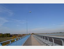 Eine Brücke mit blauem Geländer und einer asphaltierten Fahrbahn, daneben ein Gehweg. Im Hintergrund sind Bäume und eine weitläufige Landschaft unter klarem, blauem Himmel zu sehen.