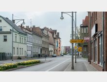 Eine städtische Straße mit mehrstöckigen Wohn- und Geschäftsgebäuden, teils aus Ziegelstein und Putzfassaden, links und rechts der Fahrbahn. Im Vordergrund befindet sich ein Mittelstreifen mit blühenden Narzissen, während Verkehrsschilder, darunter ein Wegweiser nach "Hertefeld 8 km", sichtbar sind.