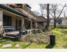 Wohnhaus mit einem Garten, das von einer überdachten Terrasse aus zugänglich ist. Die Terrasse ist mit Topfpflanzen geschmückt, während der Garten Rasenflächen, Blumenbeete und vereinzelte Bäume enthält; im Hintergrund stehen weitere Gebäude.