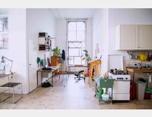 Heller Arbeitsbereich in einem geräumigen Loft mit hohen Decken und großen Fenstern. Der Raum ist teilweise als Büro mit Schreibtischen, Regalen und Pflanzen sowie als kleine Küche mit Herd, Schränken und Abwaschbereich genutzt.