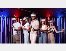 Innenraum einer VR-Arcade mit dunkler Decke, LED-Lichtleisten in Rot und Blau und einer Wand aus vertikalen Paneelen. Mehrere VR-Headsets und Hand-Controller sind im Vordergrund zu sehen, was den Raum als Virtual-Reality-Spielbereich kennzeichnet.