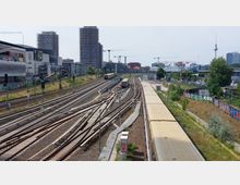 Blick auf ein weitläufiges Bahngleisfeld in einer urbanen Umgebung mit mehreren parallelen Gleisen, fahrenden Zügen und Signalanlagen. Im Hintergrund befinden sich moderne Hochhäuser, Baukräne und der Fernsehturm, zudem sind umliegende Gebäude und Graffiti-bedeckte Wände sichtbar.