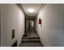 Ein schmaler, weiß gestrichener Kellerflur mit Betonboden, einer Treppenstufe in der Mitte und sichtbaren Rohren an der Decke. An der rechten Wand hängt ein roter Feuerlöscher; eine geöffnete Tür im Hintergrund führt zu einem weiteren Raum.