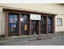 Eingangsbereich eines Gebäudes mit der Beschriftung „Robinson Tribüne“, bestehend aus mehreren gläsernen Türen und Fenstern mit braunen Rahmen. Im Inneren sind bunte Werbeplakate sichtbar, die Fassade ist cremefarben mit einem flachen Vordach.