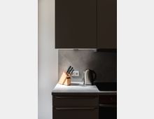 Moderne Küchenarbeitsplatte mit einer hellen Oberfläche, darauf ein Holzmesserblock mit mehreren Messern, ein Edelstahl-Wasserkocher und eine Steckdose an der Wand; der Hintergrund zeigt eine graue Wand und darüber dunkle Küchenschränke.