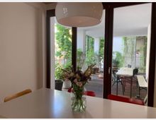 Ein heller Innenraum mit einem weißen Tisch, auf dem ein Blumenstrauß in einer Vase steht, im Vordergrund. Durch eine verglaste Tür mit dunklem Rahmen ist eine möblierte Terrasse mit Pflanzen in Töpfen und Kletterpflanzen an den Wänden zu sehen.