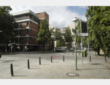 Straßenszene mit Wohngebäude aus roten Backsteinen und modernen weißen Elementen, umgeben von Bäumen. Im Vordergrund eine gepflasterte Kreuzung mit Pollern, einer Straßenlaterne und einem Straßenschild mit der Aufschrift „Goethestraße“.