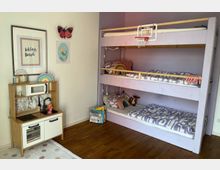 Kinderzimmer mit Etagenbett in hellem Lila, ausgestattet mit gemusterten Bettdecken und Kissen. Links steht eine kleine Spielküche aus Holz vor einer weißen Wand mit bunten Dekorationen, darunter Bilder, ein Regenbogen und ein Schmetterling.