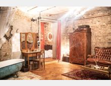 Ein Raum im Vintage-Stil mit freiliegenden Backsteinwänden, einem Holzboden und antiken Möbeln wie einem Schminktisch mit Spiegel, einem großen Holzschrank und einer Ledercouch. Im Vordergrund steht eine freistehende Badewanne, und durch einen roten Vorhang ist ein angrenzender Raum sichtbar.