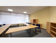 Ein einfacher Seminarraum mit gelben Wänden, einer Deckenbeleuchtung und Lamellenjalousien vor einem großen Fenster. Der Raum enthält Tische in U-Form, blaue Stühle sowie Regale und Schränke aus Holz.