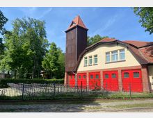 Historisches Gebäude einer Feuerwehr mit Walmdach und hohem Turm aus Holz, gelber Putzfassade, roten Ziegeltönen und markanten roten Toren im unteren Bereich. Im Vordergrund befindet sich ein eingezäunter Fahrradständer, umgeben von Bäumen in einer grünen Umgebung.