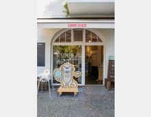 Eine kleine Eisdiele mit der Aufschrift "Gimme Gelato" über dem Eingang. Der Eingang besteht aus einer weißen Tür mit einem großen Glasfenster und abgerundeten Fensterelementen, davor steht eine auf einem Rollgestell montierte Holzfigur eines Otters mit einem Eis.