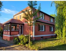 Ein zweistöckiges Wohnhaus mit orangefarbener Fassade, weißem Sockel und roten Ziegeldächern. Das Gebäude hat mehrere Fenster mit braunen Fensterläden, einen kleinen Wintergarten-Eingangsbereich sowie einen Garten mit Rasen, Bäumen und Sträuchern.