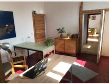 Ein heller Raum mit einfacher Büroeinrichtung, bestehend aus Schreibtischen, Stühlen und einer Holzkommode mit Zimmerpflanzen. Holztüren führen in einen weiteren Raum mit Sitzgelegenheiten, und der Boden besteht aus rotem und hellem Holz.