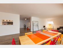 Weitläufiger Wohnraum mit hellen Wänden und Holzboden. Im Vordergrund befindet sich ein Holztisch mit bunten Stühlen, im Hintergrund eine Sitzecke mit grauem Sofa, einem kleinen Fernseher, einer Stehleuchte und einer großen Wandöffnung zur Küche.