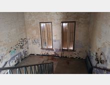 Innenraum eines stark verfallenen Treppenhauses mit abblätternder Farbe und Graffiti an den Wänden sowie einem Metallgeländer im Vordergrund. Zwei hohe Fenster mit Rahmen lassen Licht herein; der Boden ist karg und teilweise mit herumliegenden Teilen bedeckt.