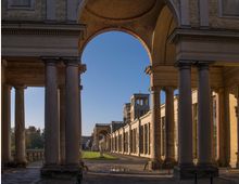 Großer Säulengang mit Rundbögen aus Sandstein in klassizistischer Architektur, der zu einem Gebäude mit zentralem Turm führt; im Hintergrund gepflegter Rasen und Bäume unter klarem Himmel.