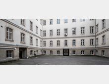 Innenhof eines historischen mehrstöckigen Gebäudes mit hellen, verputzten Wänden und zahlreichen Fenstern mit Rahmen aus Stein. Der gepflasterte Boden ist leer, und ein Durchgang führt durch die mittlere Wand.