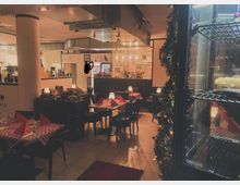 Gemütliches Restaurant mit rot-weiß karierten Tischdecken und gedämpfter Beleuchtung. Im Hintergrund befindet sich eine Bar mit Gläsern und Flaschen, daneben ist ein offener Küchenbereich sichtbar. Ein Kühlregal mit Kuchen befindet sich im Vordergrund rechts.