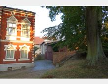 Backsteinbau mit weißen Fensterrahmen und einem Satteldach, daneben Kopfsteinpflasterweg, der zu einem Nebengebäude und Garagentoren führt. Im Vordergrund ein großer Baum und eine kleine Treppe mit Geländer, umgeben von Grünflächen.