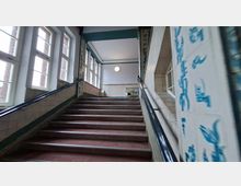 Innenansicht eines Treppenhauses mit breiter Treppe und schwarzen Handläufen, flankiert von hohen Sprossenfenstern auf der linken Seite. Die Wände sind teils gekachelt und zeigen blaue Schriftzeichen bzw. Verzierungen, oben führt die Treppe zu einem hellen Flur mit runder Deckenleuchte.