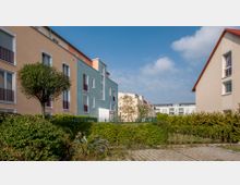 Wohngebiet mit modernen Mehrfamilienhäusern in hellen Farben, umgeben von gepflegten Hecken und Bäumen. Im Vordergrund sind zwei Fahrräder und ein gepflasterter Weg zu sehen.