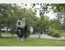 Grünfläche mit modernen Skulpturen aus Metall auf einem Betonsockel, umgeben von Bäumen und Rasen. Im Hintergrund ist eine U-Bahn-Station mit dem "U"-Symbol erkennbar.