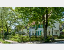 Weißes Wohnhaus mit blauen Fensterläden und Satteldach, umgeben von einem Garten mit Holzlattenzaun und blühenden Bäumen. Im Vordergrund verläuft ein Gehweg neben einem großen Baum, Straßenschilder und ein Fahrradbügel sind ebenfalls sichtbar.