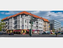 Straßenecke in einem städtischen Viertel mit einem Wohn- und Geschäftsgebäude im Vordergrund, das graue Fassaden und rote Ziegeldächer hat. Weitere Elemente sind Straßenschilder („Scharnweberstraße“ und „Kapweg“), ein Restaurant im Erdgeschoss und mehrere parkende sowie fahrende Autos, umgeben von moderner und traditioneller Architektur.