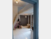 Raum mit hellem Holzboden und einer grauen Wand, eingerichtet mit einer einfachen Matratze, Kissen und einer Pflanze in einem Topf. Eine offene Holzleiter führt zu einer erhöhten Plattform, während eine hängende Lampe den Raum beleuchtet.