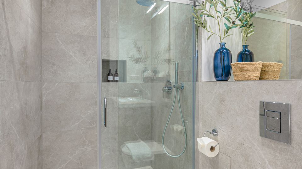 Modernes Badezimmer mit großformatigen grauen Fliesen, bodengleicher Glasdusche mit Handbrause und Ablaufrinne sowie großer Spiegelwand. Rechts sind eine wandhängende Toilette, ein Spülkastenpanel und eine Ablage mit blauen Vasen und Körben zu sehen.