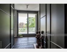 Flur mit dunklem Holzboden und modernen, schlicht gestalteten Wänden, am Ende große bodentiefe Fenster mit Blick auf eine Terrasse und grüne Bäume; am rechten Rand stehen Holzstatuen von drei stehenden Figuren.