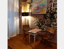 Gemütlicher Innenraum mit Holzboden, zwei Holzstühlen und einem kleinen weißen Tisch mit rotem Tischtuch. Die Wände sind mit einer Kombination aus künstlerischen Collagen, Gemälden und einem warm erleuchteten Stehlampe dekoriert, während ein heller Vorhang im Hintergrund das Fenster verdeckt.