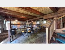 Innenraum eines rustikalen Cafés in einem umgebauten Stein- und Fachwerkgebäude mit sichtbaren Holzbalken, Kopfsteinpflasterboden und kleinen Tischen mit Stühlen. Im Raum stehen eine Theke mit Flaschen, gerahmte Bilder an den Steinwänden sowie eine Holztreppe mit Geländer im Vordergrund.