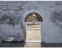 Mauer aus weißen Putzsteinen mit einem kleinen, bogenförmigen Durchgang, der von Ziegeln umrahmt ist; dahinter ist ein Hof mit einer weiteren Mauer und einer Holztür zu sehen. Im Hintergrund befinden sich Bäume.