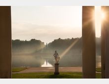 Eine Parklandschaft mit einem symmetrischen Wasserbecken in der Mitte, umgeben von Bäumen im Hintergrund. Im Vordergrund steht eine weiße Statue auf einem Sockel, eingerahmt von zwei Pfeilern, während die tiefstehende Sonne durch die Säulen scheint.
