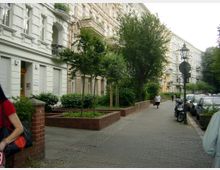 Straßenszene mit historischen Wohngebäuden im Gründerzeitstil, verziert mit Balkonen und Stuckelementen. Ein Gehweg mit Bäumen, Straßenlaternen und geparkten Fahrzeugen verläuft entlang der Gebäude; im Vordergrund befinden sich kleine begrünte Flächen mit niedrigen Backsteinmauern.